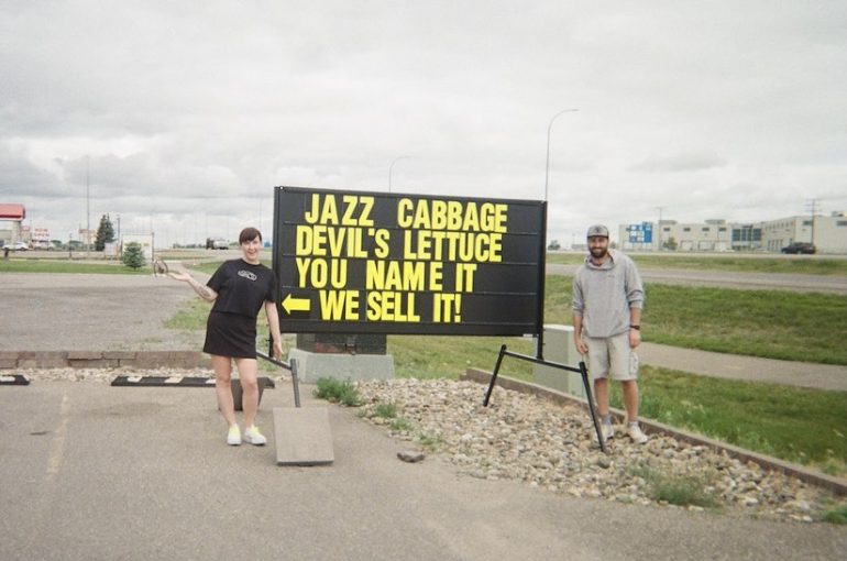 Two people standing beside a roadside sign that reads: JAZZ CABBAGE, DEVIL'S LETTUCE, YOU NAME IT, WE SELL IT"