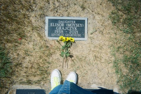 A woman's feet at the grave of her grandmother with yellow flowers laying on top of the gravestone.