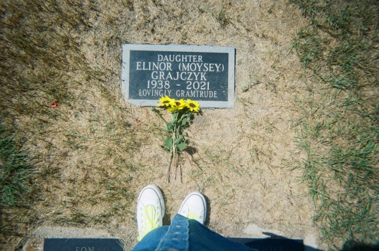 A woman's feet at the grave of her grandmother with yellow flowers laying on top of the gravestone.
